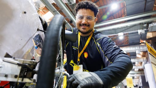 Homem usando óculos de proteção e luvas trabalha na manutenção de um veículo de competição, ao lado de uma das rodas, em ambiente de oficina.