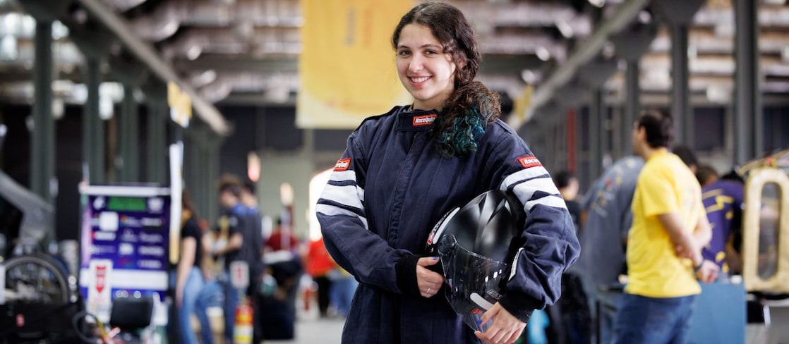Jovem pilota sorri para a câmera usando macacão de corrida azul e segurando um capacete preto. Ao fundo, outras pessoas e veículos estão em uma área de boxes de uma competição automobilística. 
