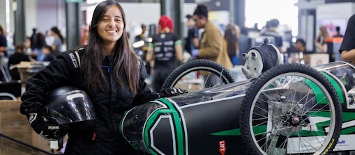 Jovem pilota sorri para a câmera usando macacão de corrida preto e segurando um capacete. Ao seu lado, está um veículo de competição preto e verde, com rodas estreitas e estrutura aerodinâmica, em uma área de boxes movimentada.