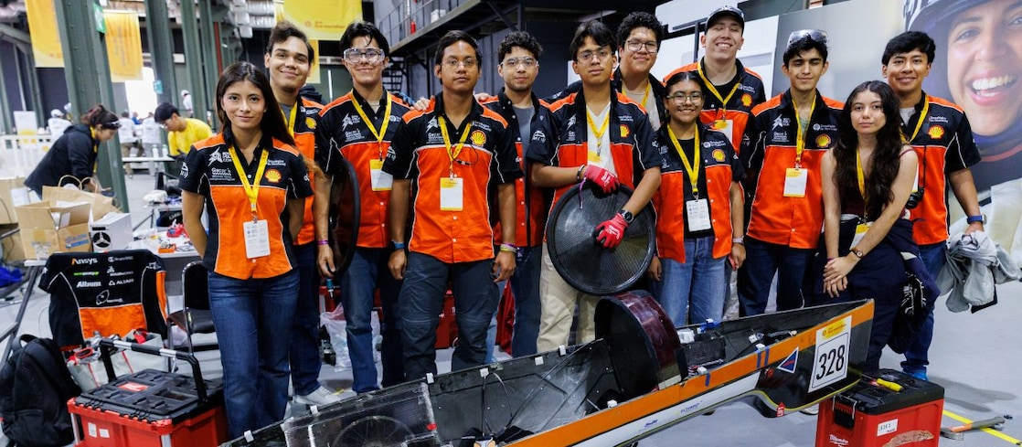 Equipe da Shell Eco-marathon posa em uma área de boxes ao lado de um veículo de eficiência energética preto e laranja, identificado com o número 328. Os integrantes usam uniformes nas mesmas cores, com credenciais do evento, enquanto ferramentas e equipamentos de trabalho aparecem ao redor do veículo.