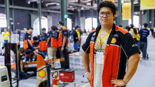 Grupo de participantes da Shell Eco-marathon posa ao lado de um veículo de eficiência energética preto, identificado com o número 330 e o logotipo da Shell. A equipe, formada por dez pessoas, veste majoritariamente camisetas amarelas e equipamentos de segurança. A foto foi registrada em uma área interna de competição, com boxes e estruturas técnicas ao fundo.