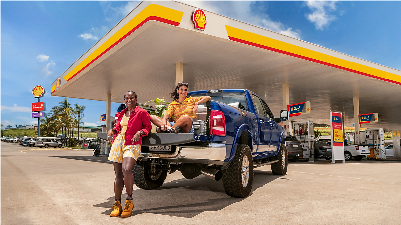 Carro estacionado em um posto Shell e duas mulheres, uma à frente do carro e outra na carroceria do carro