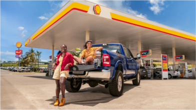 Carro estacionado em um posto Shell e duas mulheres, uma à frente do carro e outra na carroceria do carro