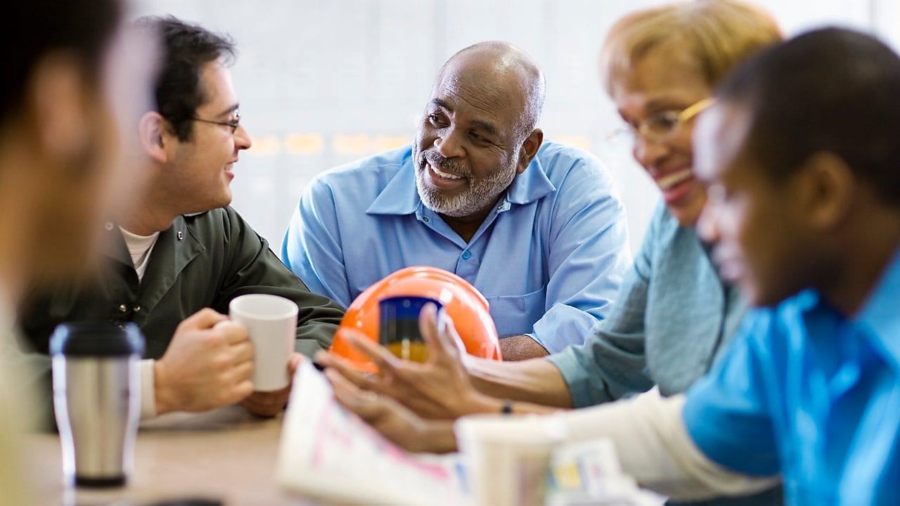 Cinco pessoas conversam ao redor de uma mesa de escritório, em clima de descontração