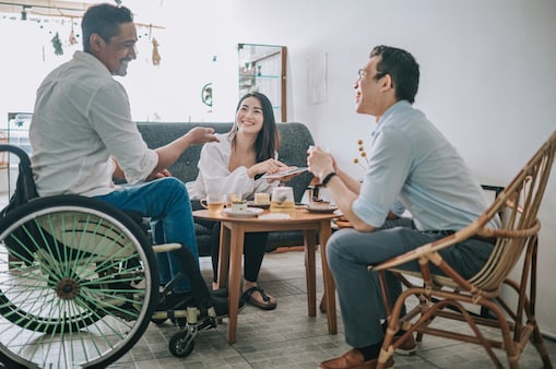 Três pessoas conversando em um ambiente acolhedor, incluindo um homem em cadeira de rodas, uma mulher sorrindo e outro homem sentado, simbolizando diversidade e inclusão.