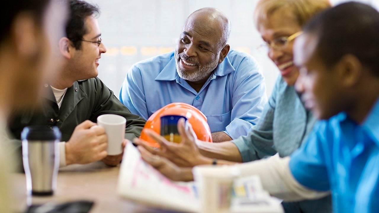 Cinco pessoas conversam ao redor de uma mesa de escritório, em clima de descontração