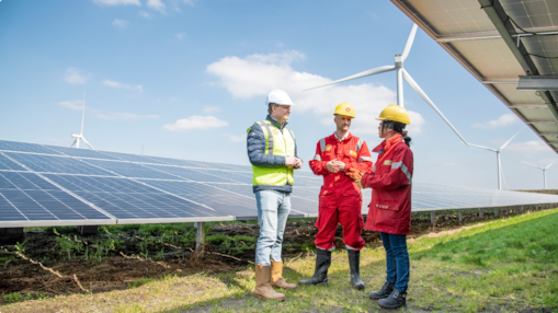 Pessoas usando equipamento de segurança conversam em frente a painéis solares e turbinas eólicas