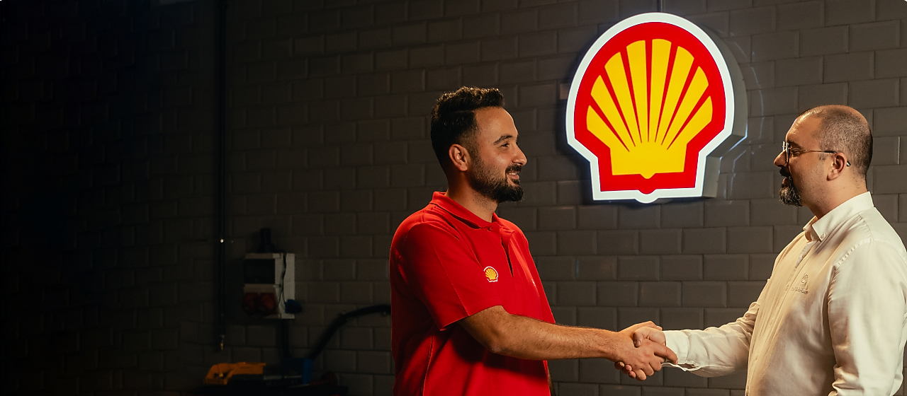 Dois homens, um deles com uma blusa vermelha e com a concha Shell, apertam as mãos