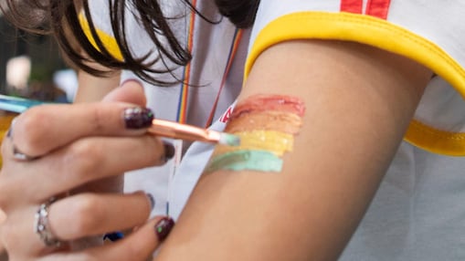 Uma pessoa pinta, em seu braço, a bandeira com as cores do arco-íris: vermelho , laranja, amarelo, verde, azul e roxo.
