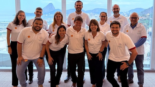 Colaboradores da Shell Brasil posam sorridentes em grupo durante o encontro da rede de afinidade Generations no escritório do Rio de Janeiro. Todos vestem camisetas brancas com o logo da Shell e da rede Generations, com o Pão de Açúcar ao fundo visto pelas janelas panorâmicas.