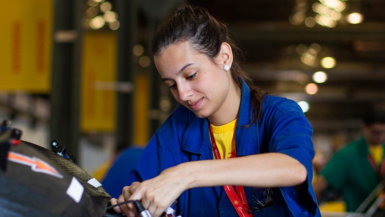 Em um fundo amarelo, vemos três ilustrações de mulheres com uniforme da Shell posicionadas lado a lado. A primeira está com um jaleco vermelho e fones de proteção, a segunda com o uniforme branco e a terceira com uniforme vermelho, fone de proteção e capacete de proteção branco.