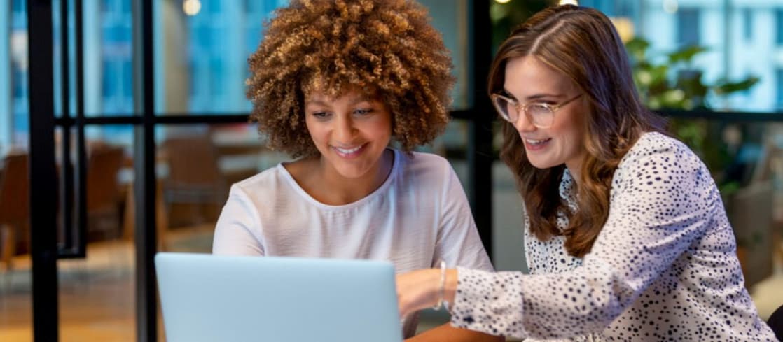 Duas mulheres sentadas lado a lado em um ambiente de escritório moderno, olhando para a tela de um laptop sobre a mesa. Uma delas aponta para a tela enquanto a outra acompanha, com uma xícara de café ao lado. 