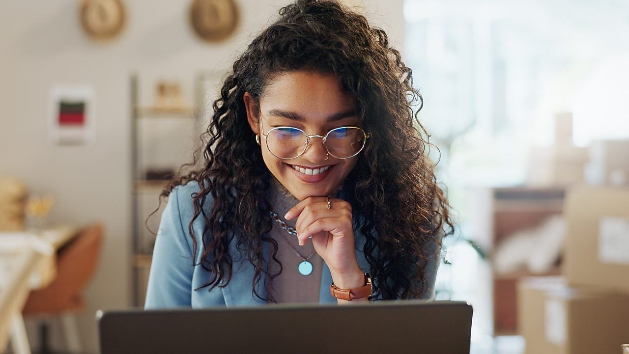 Mulher sentada em ambiente interno, com um laptop aberto sobre a mesa, olhando para a tela. Ao fundo, há iluminação natural e elementos simples do ambiente. 