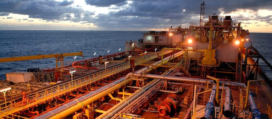 Plataforma de petróleo offshore ao entardecer, com diversas tubulações, estruturas metálicas e iluminação artificial, tendo o mar e o céu nublado ao fundo. 