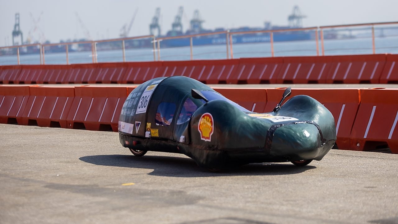 Carro protótipo aerodinâmico da Shell Eco-marathon, de cor verde escura e número 306, circulando em pista asfaltada próxima ao mar, com barreiras de segurança laranja e guindastes portuários ao fundo.