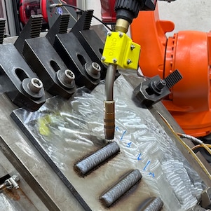  Supefície onde o metal é depositado com auxílio de um robô, usando um arame consumível  combinado com um arco elétrico que gera calor Supefície onde o metal é depositado com auxílio de um robô, usando um arame consumível combinado com um arco elétrico que gera calor