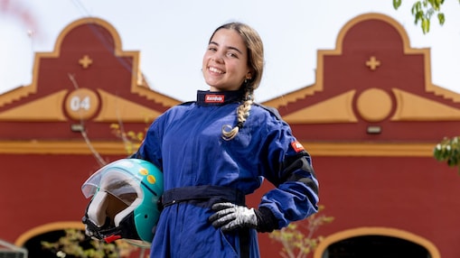 Jovem participante da Shell Eco-marathon sorri para a câmera usando macacão de corrida azul e luvas, segurando um capacete verde claro com adesivos. Ao fundo, uma construção histórica de fachada vermelha e detalhes amarelos completa a cena ao ar livre.