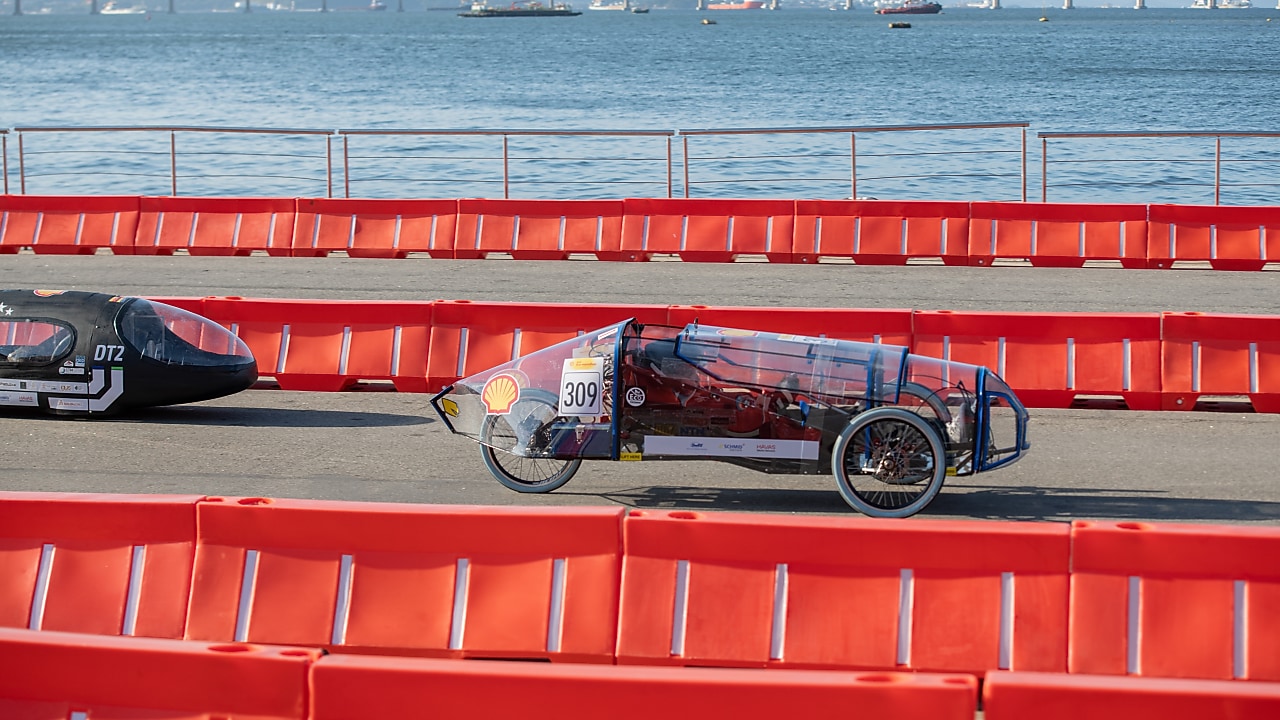 Protótipo de veículo da Shell Eco-marathon em movimento durante a competição no Rio de Janeiro. O carro, de estrutura leve e transparente, com o número 309, percorre a pista cercada por barreiras de segurança vermelhas, com o mar e embarcações ao fundo.