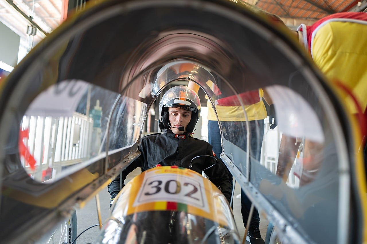 Piloto sentado dentro de um protótipo aerodinâmico com número 302, usado em competição de eficiência energética. Ele veste macacão preto e capacete com viseira, enquanto é observado por integrantes da equipe usando camisetas amarelas e vermelhas.