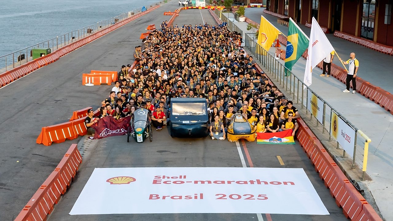 Foto oficial com centenas de participantes reunidos ao lado de protótipos veiculares na pista da Shell Eco-marathon Brasil 2025, com bandeiras do Brasil, Shell e outros parceiros ao fundo, à beira da Baía de Guanabara, no Rio de Janeiro.