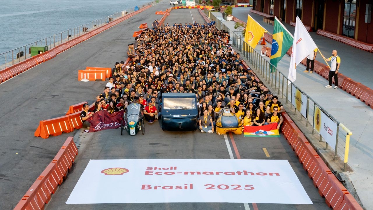 Foto oficial com centenas de participantes reunidos ao lado de protótipos veiculares na pista da Shell Eco-marathon Brasil 2025, com bandeiras do Brasil, Shell e outros parceiros ao fundo, à beira da Baía de Guanabara, no Rio de Janeiro. 