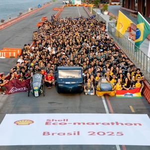 Foto oficial com centenas de participantes reunidos ao lado de protótipos veiculares na pista da Shell Eco-marathon Brasil 2025, com bandeiras do Brasil, Shell e outros parceiros ao fundo, à beira da Baía de Guanabara, no Rio de Janeiro. 