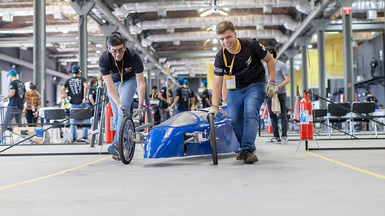 Dois estudantes empurram um protótipo aerodinâmico azul de eficiência energética em um galpão industrial durante a Shell Eco-marathon, com outros participantes e equipes ao fundo. 