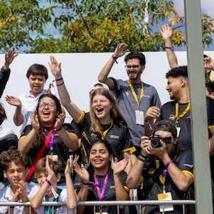 Público formado por estudantes e visitantes vibra nas arquibancadas durante a Shell Eco-marathon, com aplausos, sorrisos e registros com celular e câmera fotográfica. 