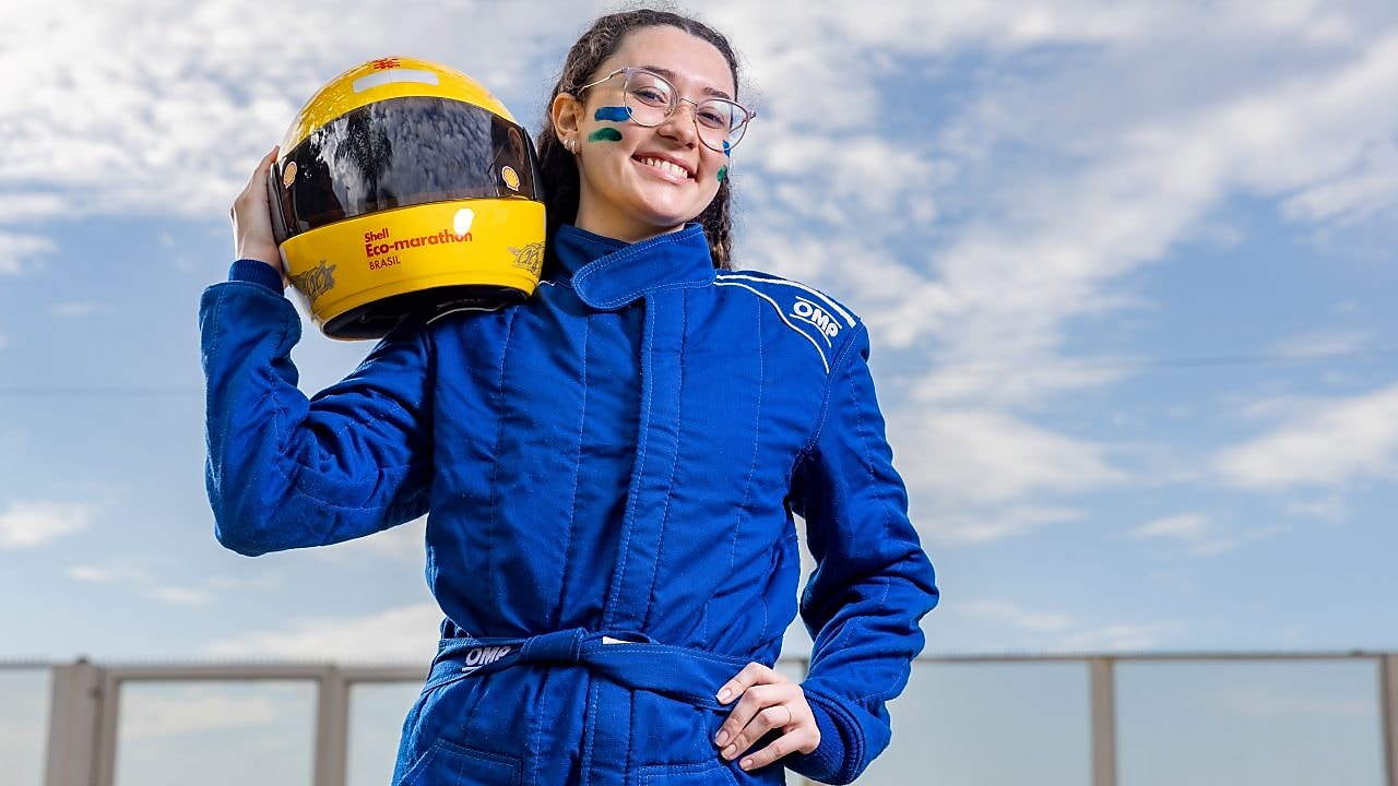 Jovem piloto sorri em pose confiante usando macacão azul e segurando capacete amarelo com o logo da Shell Eco-marathon Brasil, sob um céu azul com nuvens ao fundo. 
