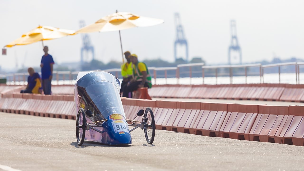 Protótipo aerodinâmico com número 314 circula na pista ao ar livre durante a Shell Eco-marathon, com barreiras de segurança ao redor e pessoas observando sob guarda-sóis ao fundo.