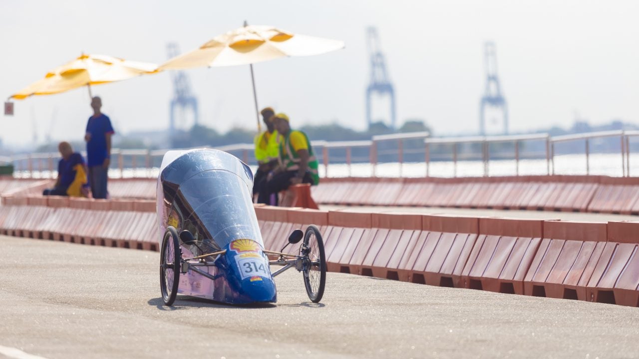 Protótipo aerodinâmico com número 314 circula na pista ao ar livre durante a Shell Eco-marathon, com barreiras de segurança ao redor e pessoas observando sob guarda-sóis ao fundo. 