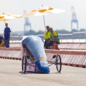 Protótipo aerodinâmico com número 314 circula na pista ao ar livre durante a Shell Eco-marathon, com barreiras de segurança ao redor e pessoas observando sob guarda-sóis ao fundo. 