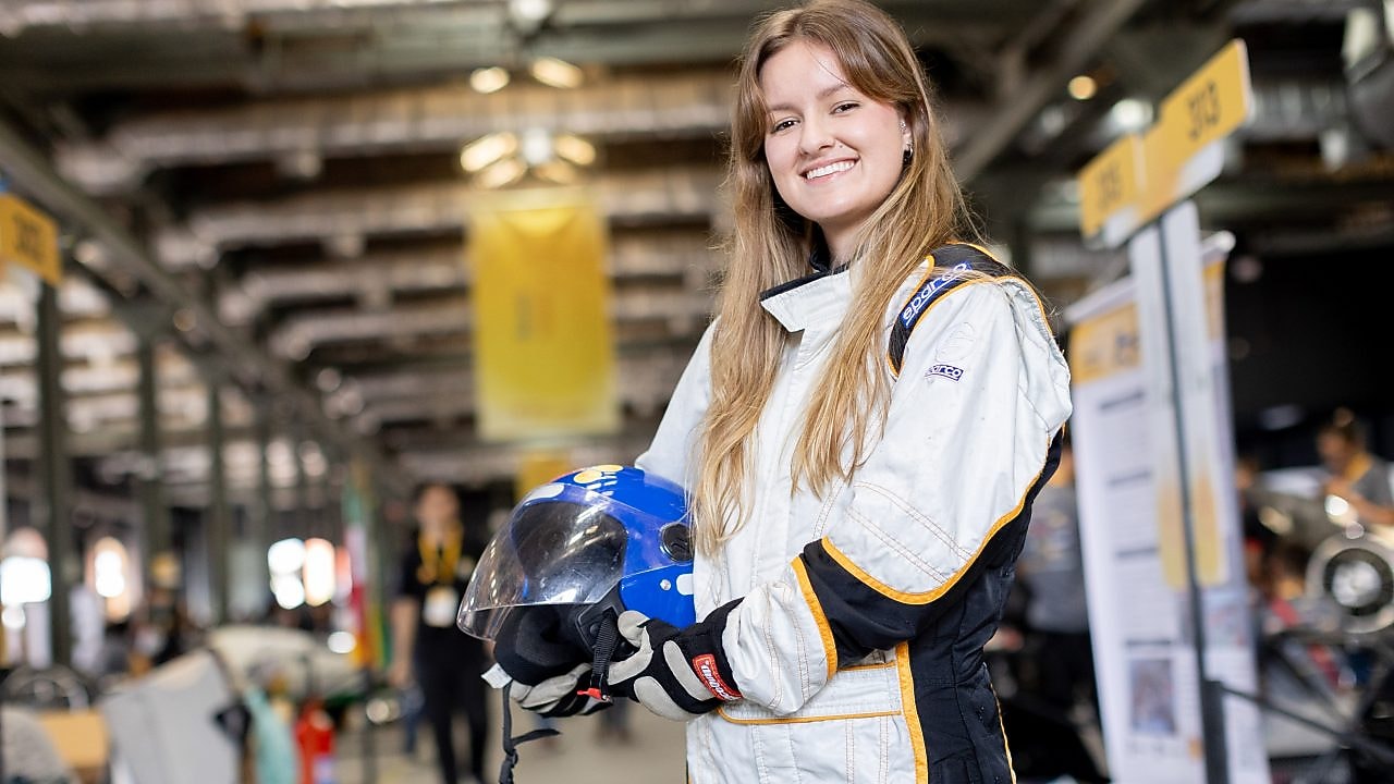 Estudante sorridente posa com macacão de corrida e capacete em mãos durante a Shell Eco-marathon, em área interna do evento com boxes de equipes ao fundo.