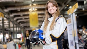 Estudante sorridente posa com macacão de corrida e capacete em mãos durante a Shell Eco-marathon, em área interna do evento com boxes de equipes ao fundo. 
