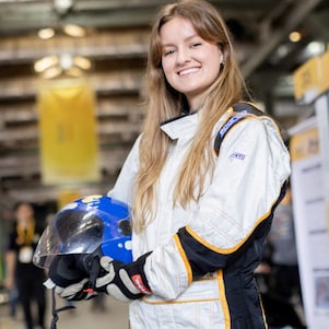 Estudante sorridente posa com macacão de corrida e capacete em mãos durante a Shell Eco-marathon, em área interna do evento com boxes de equipes ao fundo. 
