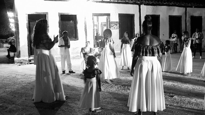 Grupo de pessoas em trajes tradicionais participando de uma apresentação cultural à noite em frente a uma casa colonial iluminada