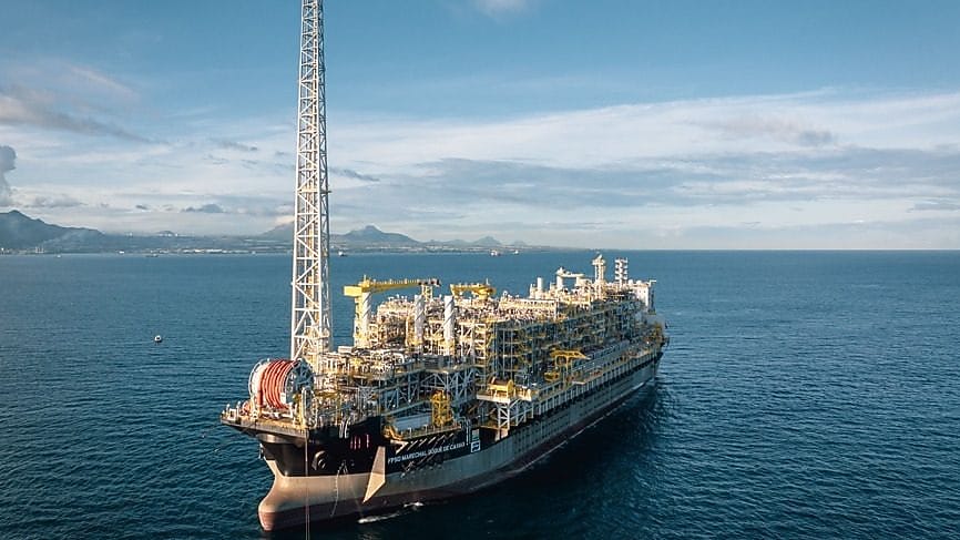 Vista aérea de uma plataforma flutuante de produção de petróleo, o FPSO Marechal Duque de Caxias, localizada no oceano. A estrutura é grande e complexa, com várias instalações, tubulações e equipamentos industriais em tons de cinza e amarelo. Ao fundo, é possível ver uma faixa de terra com montanhas ao longe, sob um céu parcialmente nublado.