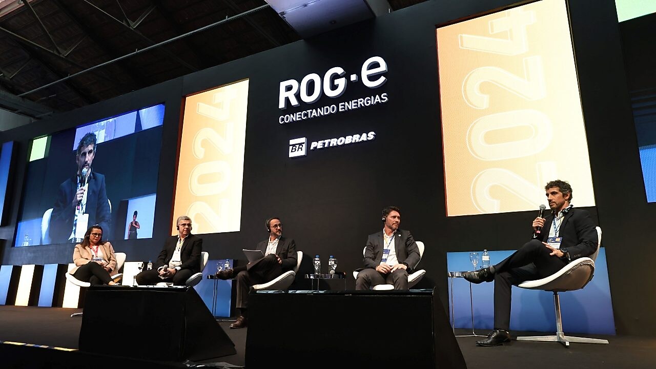 Cinco palestrantes sentados em um palco durante um painel no evento ROG.e 2024, com o logotipo da Petrobras ao fundo. O painelista à direita fala ao microfone, enquanto os outros quatro participantes escutam atentamente, todos usando fones de ouvido para tradução simultânea.
