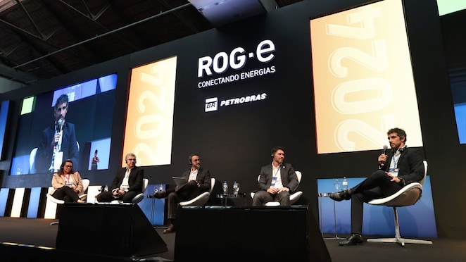 Cinco palestrantes sentados em um palco durante um painel no evento ROG.e 2024, com o logotipo da Petrobras ao fundo. O painelista à direita fala ao microfone, enquanto os outros quatro participantes escutam atentamente, todos usando fones de ouvido para tradução simultânea.