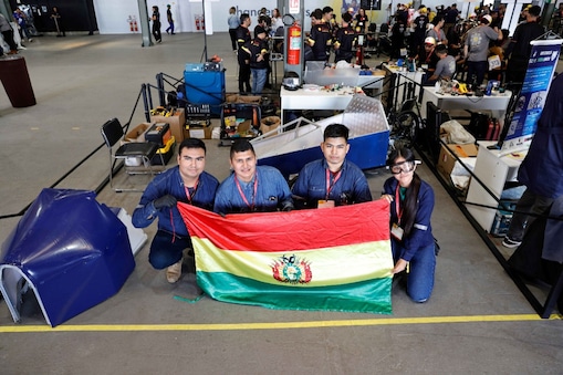 A imagem mostra a equipe Capitán Victor Ustariz da Escuela Militar de Ingeniería de Santa Cruz de La Sierra, Bolívia, durante a Shell Eco-marathon 2024 no Rio de Janeiro. Os integrantes da equipe, em uniformes azul escuro, posam com uma bandeira boliviana e um protótipo de veículo aerodinâmico azul. O ambiente é um espaço de evento com outras equipes e áreas de trabalho ao fundo.