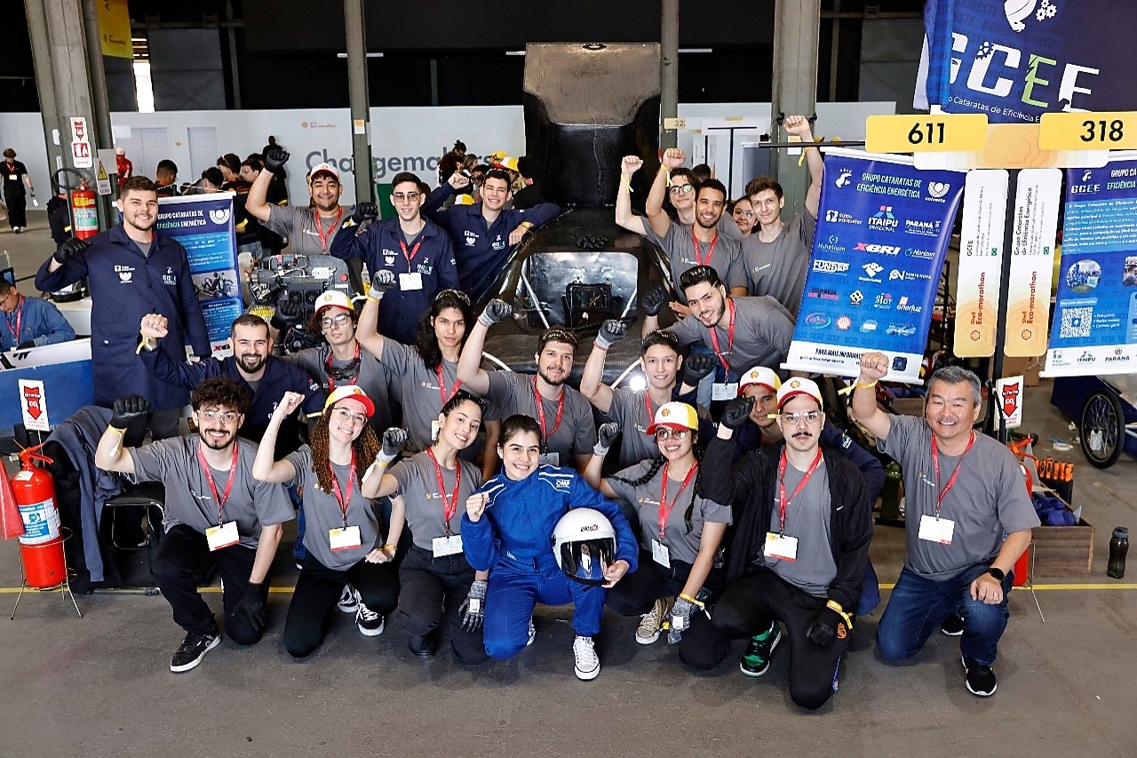 A imagem mostra o Grupo Cataratas de Eficiência Energética (GCEE) da Unioeste Foz do Iguaçu durante a Shell Eco-marathon 2024 no Rio de Janeiro. Os integrantes da equipe, em uniformes com camisetas cinza e calças escuras, tem a pilota vestida de azul e com um capacete à frente, no centro, e posam à frente de um protótipo de veículo aerodinâmico branco e azul. O ambiente é um espaço de evento com outras equipes e áreas de trabalho ao fundo.