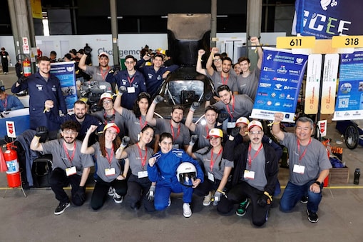 A imagem mostra o Grupo Cataratas de Eficiência Energética (GCEE) da Unioeste Foz do Iguaçu durante a Shell Eco-marathon 2024 no Rio de Janeiro. Os integrantes da equipe, em uniformes com camisetas cinza e calças escuras, tem a pilota vestida de azul e com um capacete à frente, no centro, e posam à frente de um protótipo de veículo aerodinâmico branco e azul. O ambiente é um espaço de evento com outras equipes e áreas de trabalho ao fundo.