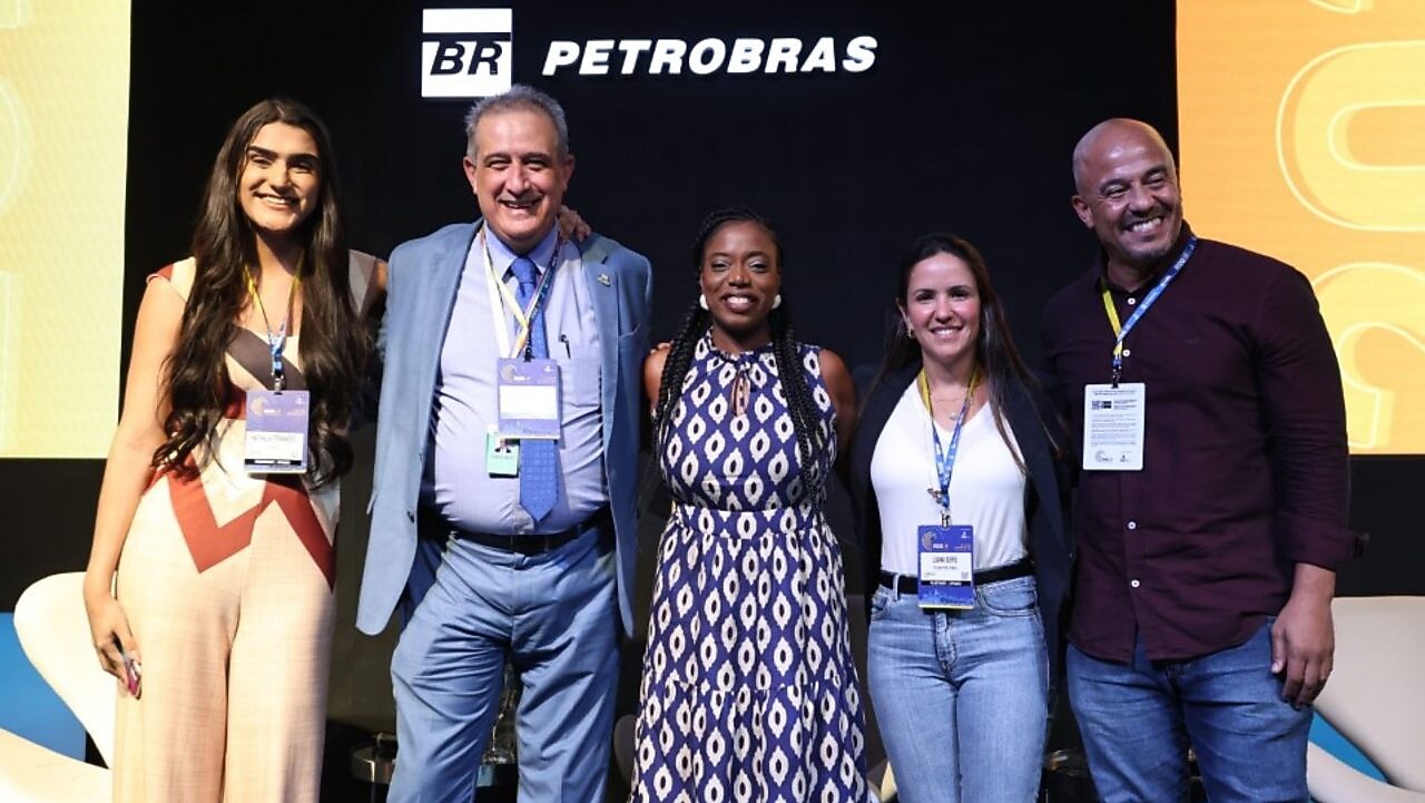 Os cinco palestrantes posam juntos no palco. Da esquerda para a direita, uma mulher jovem com cabelos longos e roupa clara, um homem mais velho de terno azul claro e gravata, uma mulher negra com vestido estampado de azul e branco, outra mulher com blusa branca e jeans, e um homem careca com camisa escura e jeans. Eles estão sorrindo, em frente a um fundo preto com o logotipo da Petrobras.