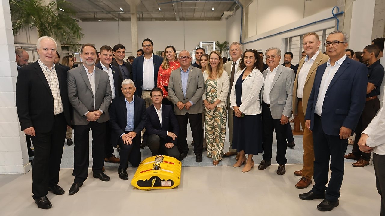 Grupo de pessoas em trajes formais, posando em ambiente interno com teto industrial e dispositivo amarelo no chão ao centro