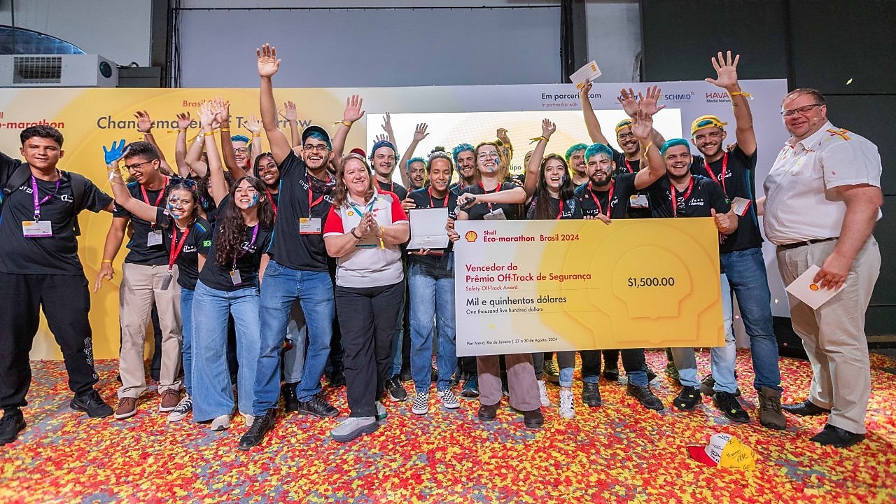 Grupo de pessoas celebra, durante o último dia do Shell Eco-Marathon Brasil 2024, com o cheque em tamanho grande de 1.500 dólares, prêmio para o ‘Vencedor do Prêmio Off-Track de Segurança”. Algumas pessoas do grupo estão com as mãos levantadas. Confetes estão espalhados pelo chão, sugerindo uma atmosfera festiva. Um homem à direita segura um microfone. Ao fundo, há um banner.