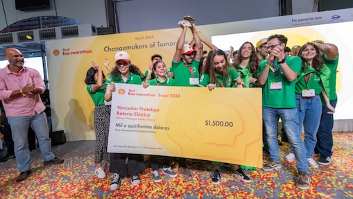 Grupo de pessoas celebra, durante o último dia do Shell Eco-Marathon Brasil 2024, com o cheque em tamanho grande de 1.500 dólares, prêmio para o ‘Vencedor Protótipo Bateria Elétrica’. Os participantes do grupo estão vestindo camisetas verdes e segurando o cheque juntos. O cenário é interno, com um carpete colorido e um banner ao fundo com os logos do Shell Eco-Marathon e ‘Changemakers of Tomorrow’.