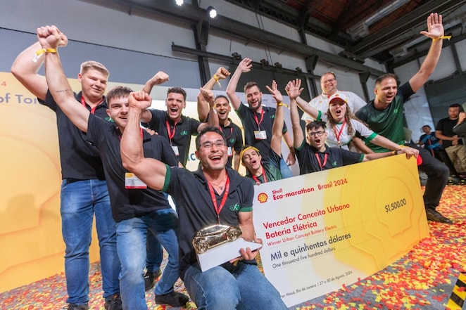 A imagem mostra um grupo de pessoas em um palco comemorando uma vitória. Eles estão com os braços levantados em triunfo, e uma pessoa está segurando um troféu. Há confetes no chão, indicando que o evento acabou de terminar ou está em modo de celebração. Um grande cheque é exibido na frente do grupo, sugerindo que eles ganharam um prêmio. O texto no cheque fornece informações sobre o evento e o prêmio: “Shell Eco-marathon Brasil 2024,” “Vencedor Categoria Urbano Bateria Elétrica,” e “$1,500.” Isso indica que esta equipe venceu na categoria de bateria elétrica urbana no Shell Eco-marathon Brasil 2014 com um prêmio de mil e quinhentos dólares.