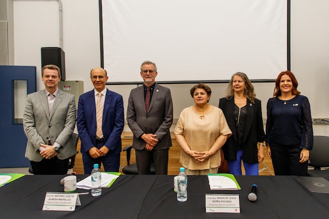Seis pessoas, três homens e três mulheres, lado a lado em trajes formais em frente a uma mesa com microfone, placas de identificação e copos de água, em um ambiente de conferência com uma tela de projeção ao fundo.