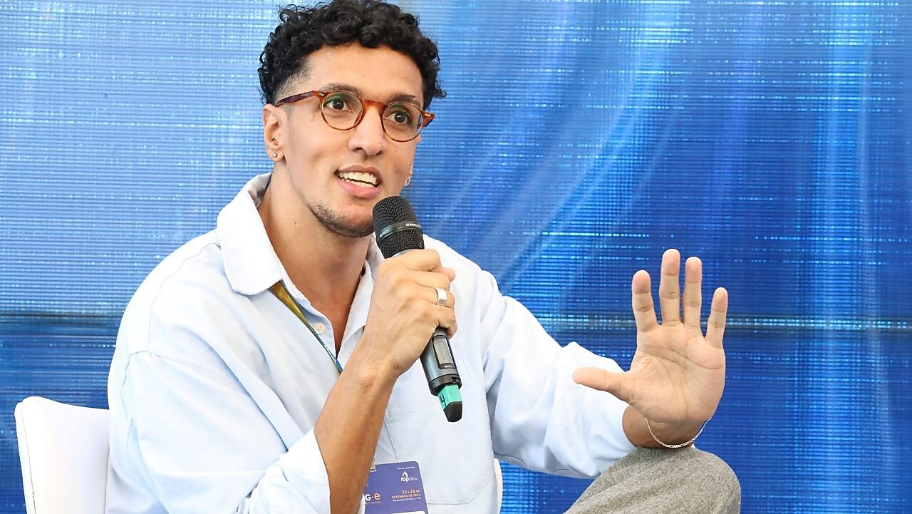Homem sentado em uma cadeira, segurando um microfone com a mão direita enquanto gesticula com a mão esquerda. Ele usa óculos de armação redonda, camisa clara e calças cinzas, com um crachá pendurado no pescoço que contém o nome 'Felipe Barbosa' e o logotipo da Shell ao fundo. O fundo da imagem é azul, e o homem parece estar falando em um painel ou evento.