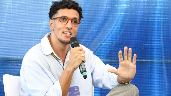 Homem sentado em uma cadeira, segurando um microfone com a mão direita enquanto gesticula com a mão esquerda. Ele usa óculos de armação redonda, camisa clara e calças cinzas, com um crachá pendurado no pescoço que contém o nome 'Felipe Barbosa' e o logotipo da Shell ao fundo. O fundo da imagem é azul, e o homem parece estar falando em um painel ou evento.